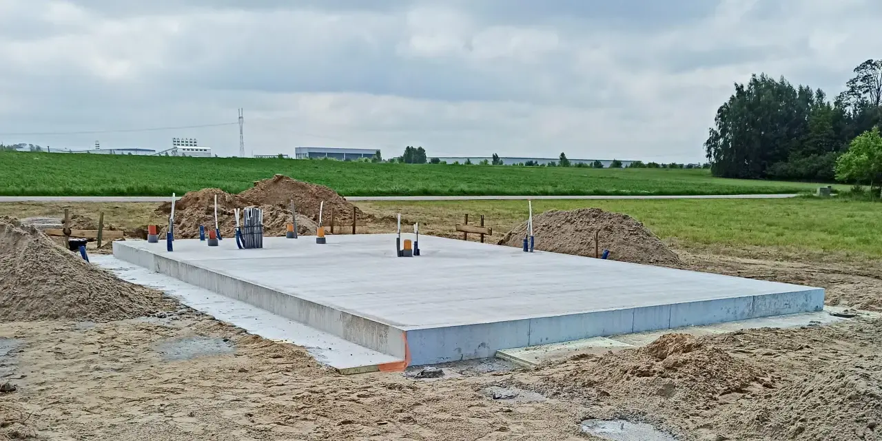 Gotowa płyta fundamentowa na budowie, otoczona piaskiem i zielenią. W tle widać pola i budynki przemysłowe.