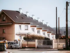 Rząd jasnych domów szeregowych z ciemnymi dachami i białymi kominami. Przed jednym z domów stoi srebrny samochód.